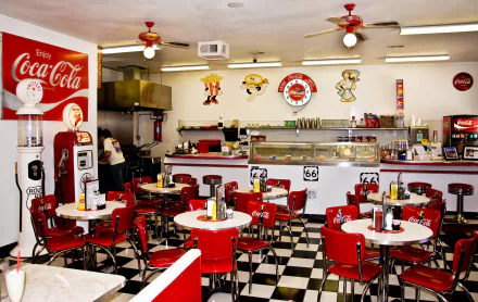 Vintage diner interior with red chairs and white tables, classic Coca-Cola decor, checkered floor, ceiling fans, and retro restaurant elements in 4K Ultra HD.
