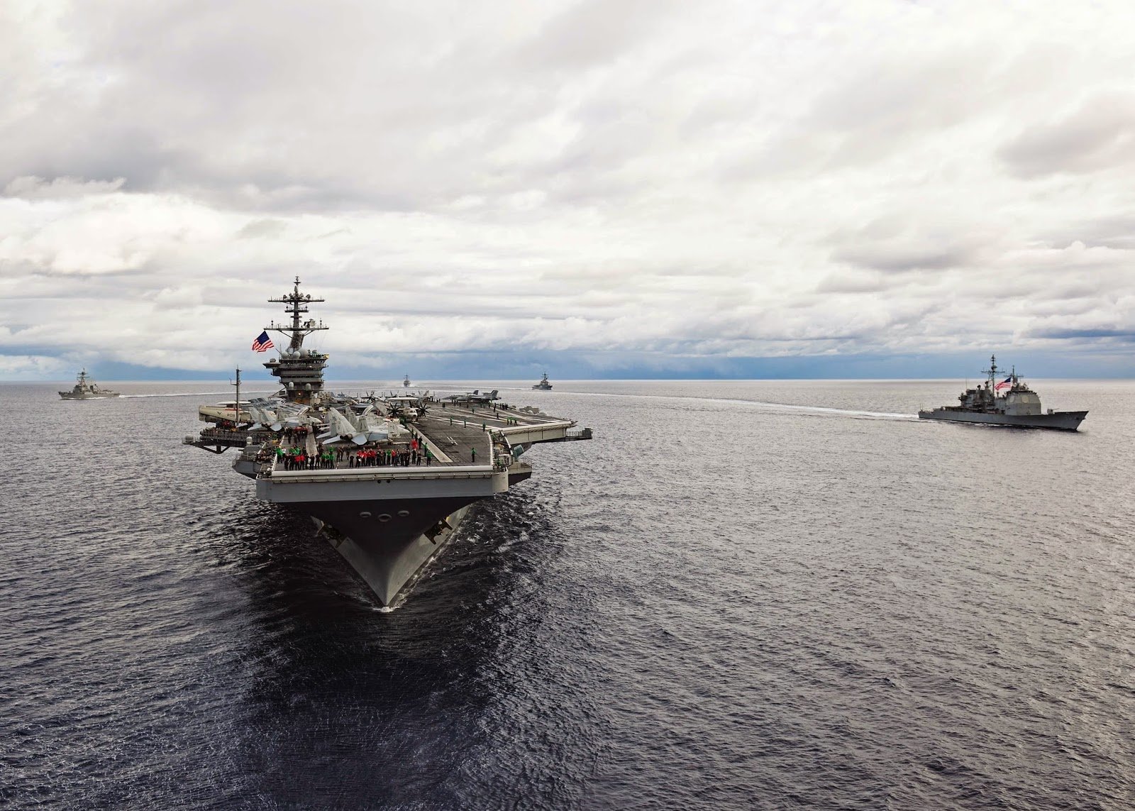 HD desktop wallpaper of the USS Theodore Roosevelt (CVN-71) aircraft carrier sailing in the ocean with accompanying navy warships under a cloudy sky.
