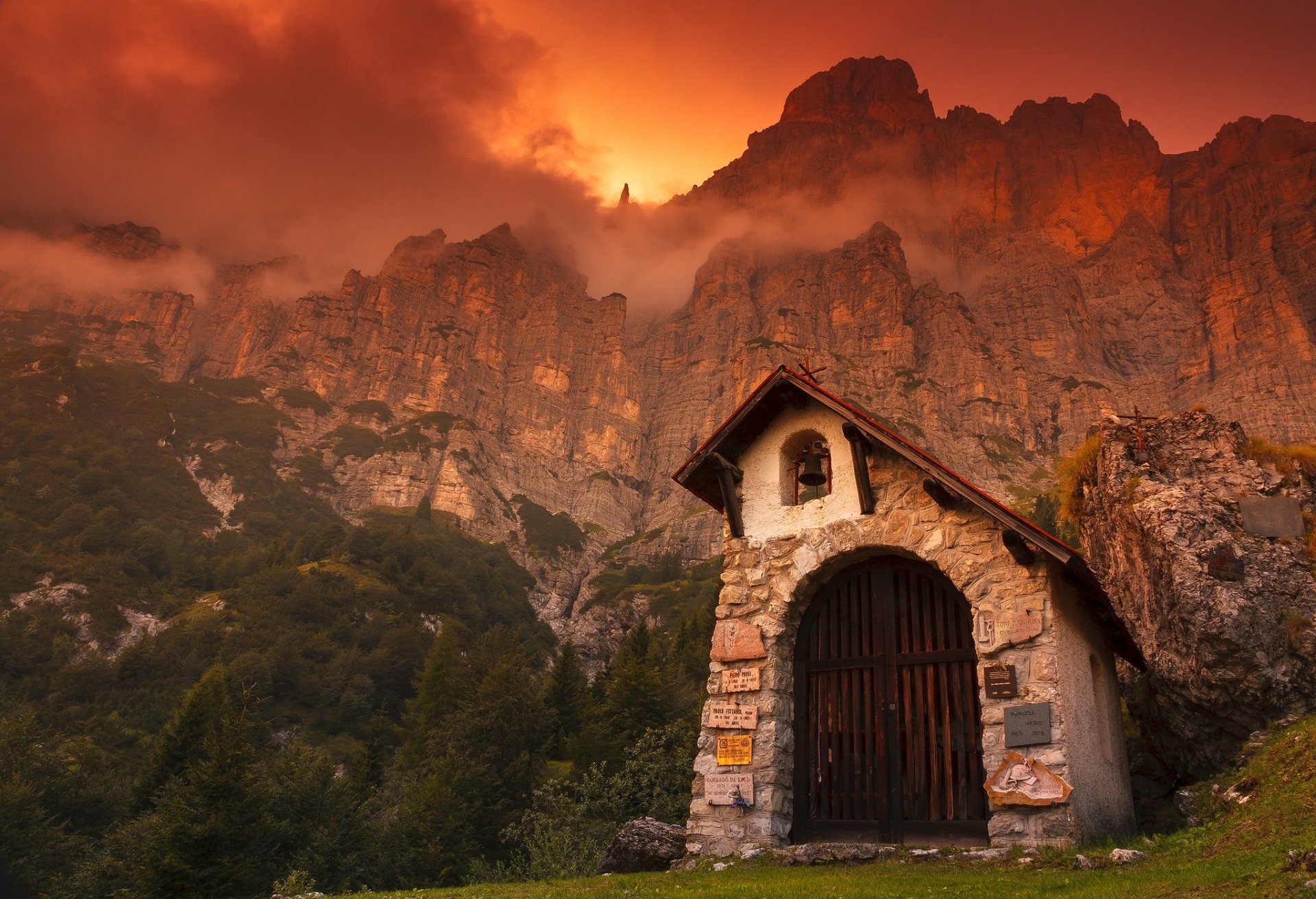 Stone chapel on a cliff at sunset in the Dolomites, Italy, with dramatic mountain peaks glowing under a fiery sky.