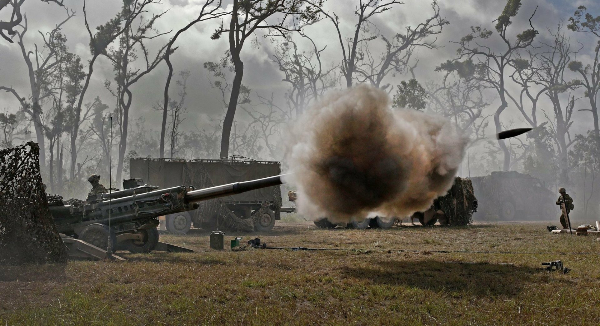 HD desktop wallpaper featuring a military M777 howitzer artillery weapon firing amid a smoky, barren landscape with leafless trees in the background.
