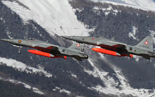 Two military Northrop F-5 jet fighters with red external tanks fly over snow-covered mountains — 2K Quad HD PC desktop wallpaper/background of warplane aircraft.