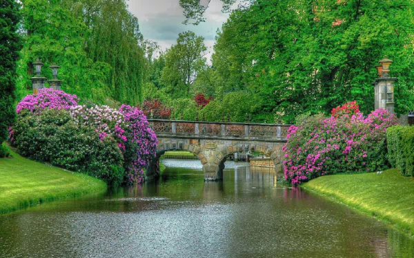 A serene spring garden with a stone bridge over a pond, surrounded by lush trees, blooming bushes, and colorful flowers in a peaceful park setting.