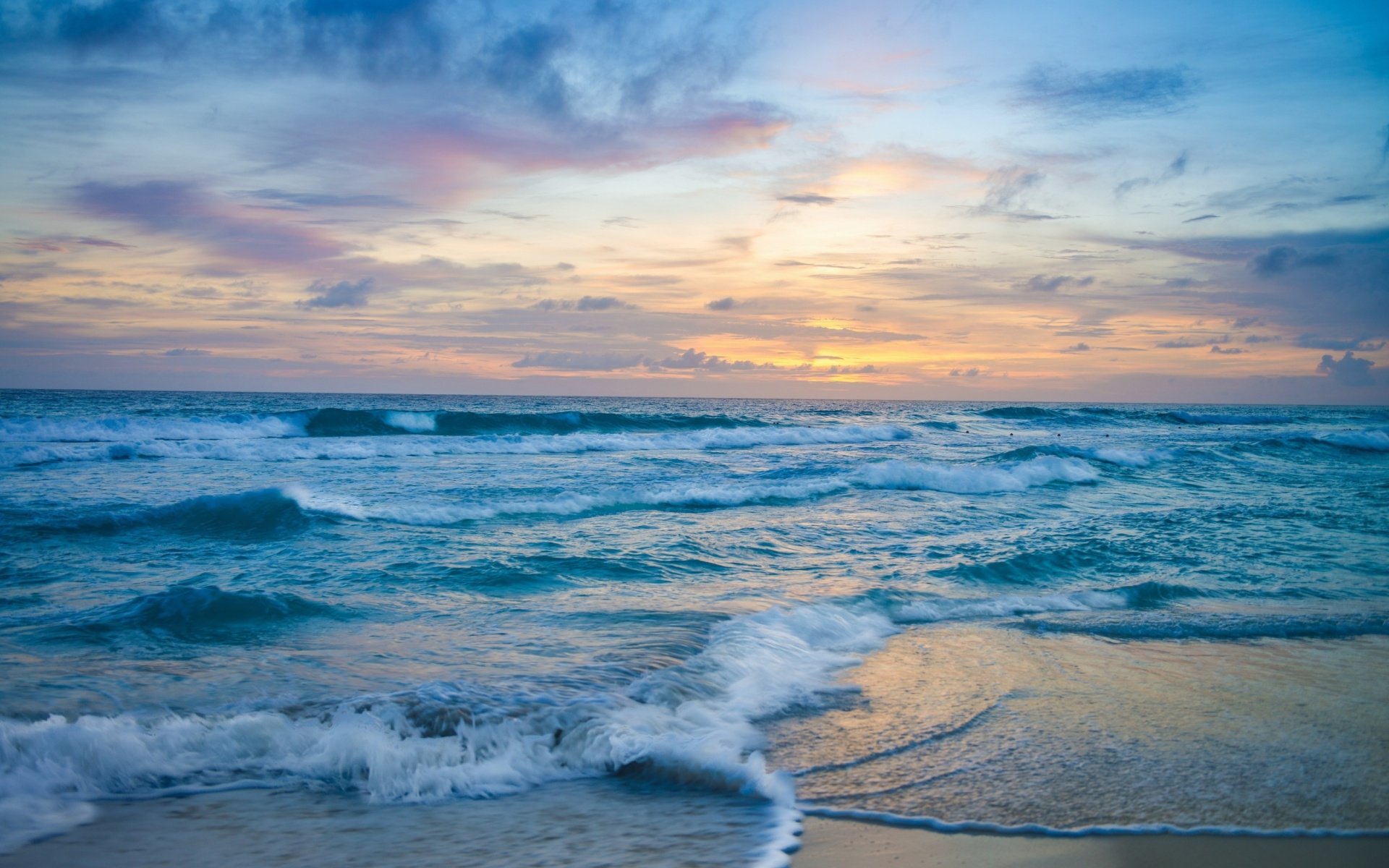 HD desktop wallpaper of a blue ocean wave gently washing ashore under a colorful sunset sky on the horizon, showcasing nature's serene beauty.