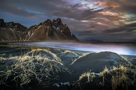 Dramatic 4K Ultra HD landscape of Vestrahorn mountain with clouds above, ocean waves, and sandy terrain illuminated under moody lighting.