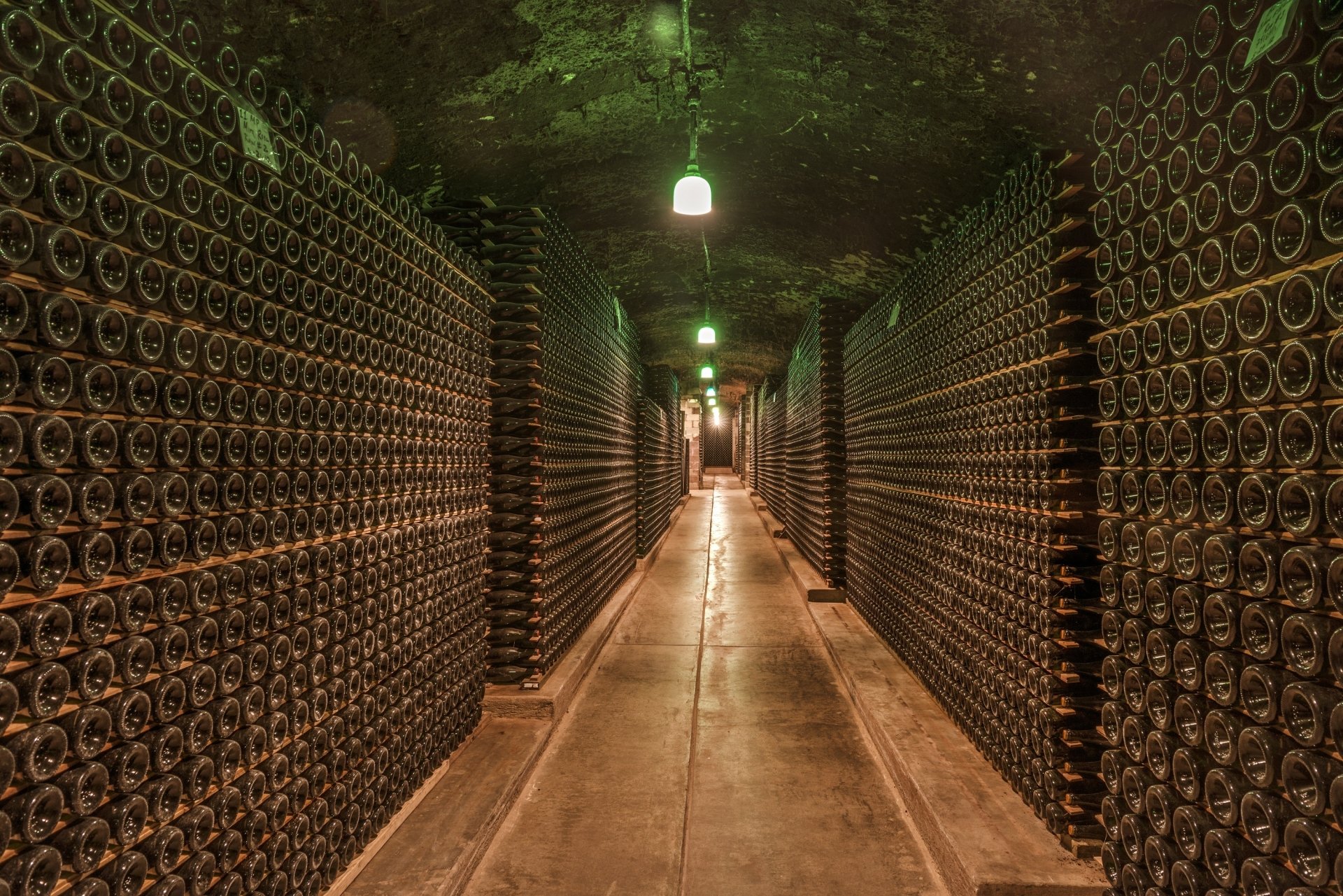 2K Quad HD PC desktop wallpaper: underground, man-made wine cellar corridor lined floor-to-ceiling with stacked bottles under green pendant lights.