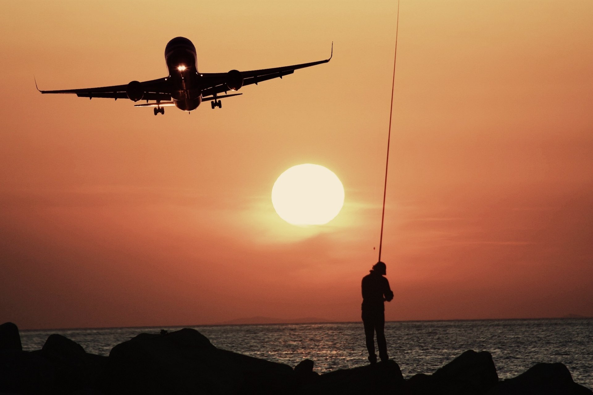 A silhouette of a fisherman casting a line as an aircraft descends against a vibrant sunset horizon, capturing the essence of evening sports photography by the water.