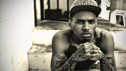A black-and-white HD desktop wallpaper featuring Kid Ink, a tattooed artist wearing a RECKLESS cap, sitting with his chin resting on clasped hands, exuding a thoughtful demeanor.