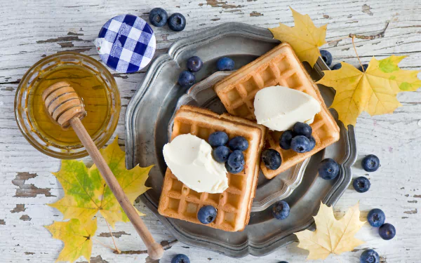 HD desktop wallpaper featuring blueberry-topped waffles with honey and ice cream, surrounded by fresh blueberries and autumn leaves on a rustic wooden table.