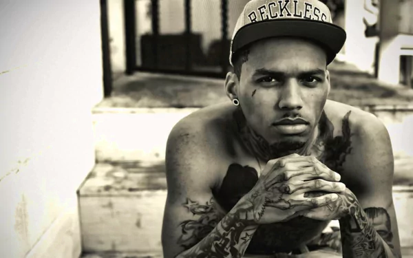 A black-and-white HD desktop wallpaper featuring Kid Ink, a tattooed artist wearing a RECKLESS cap, sitting with his chin resting on clasped hands, exuding a thoughtful demeanor.