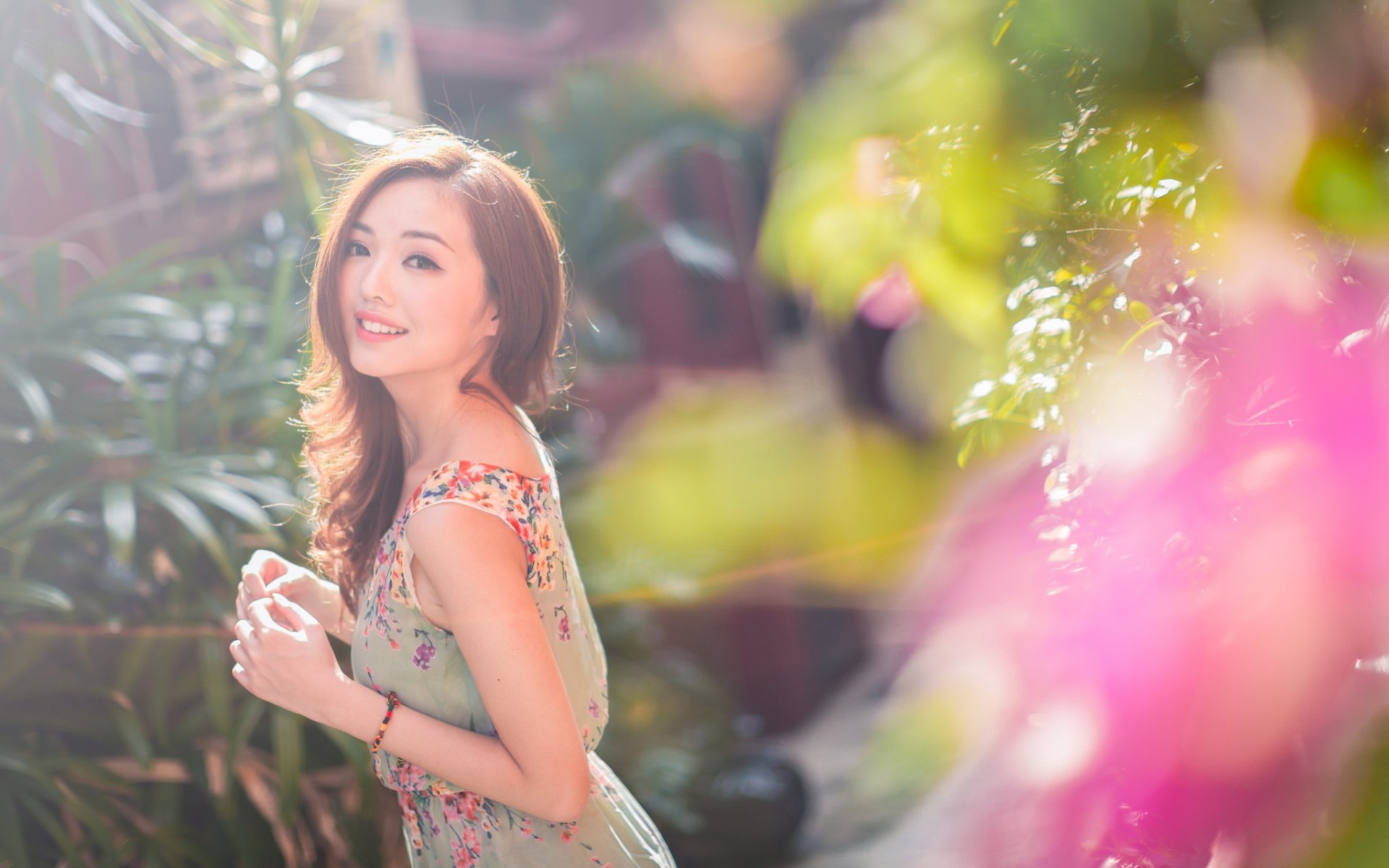 A smiling brunette woman with brown eyes stands outdoors in a sunny setting, surrounded by vibrant, bokeh-enhanced flora. The Asian model's serene demeanor is captured in this high-definition desktop wallpaper.