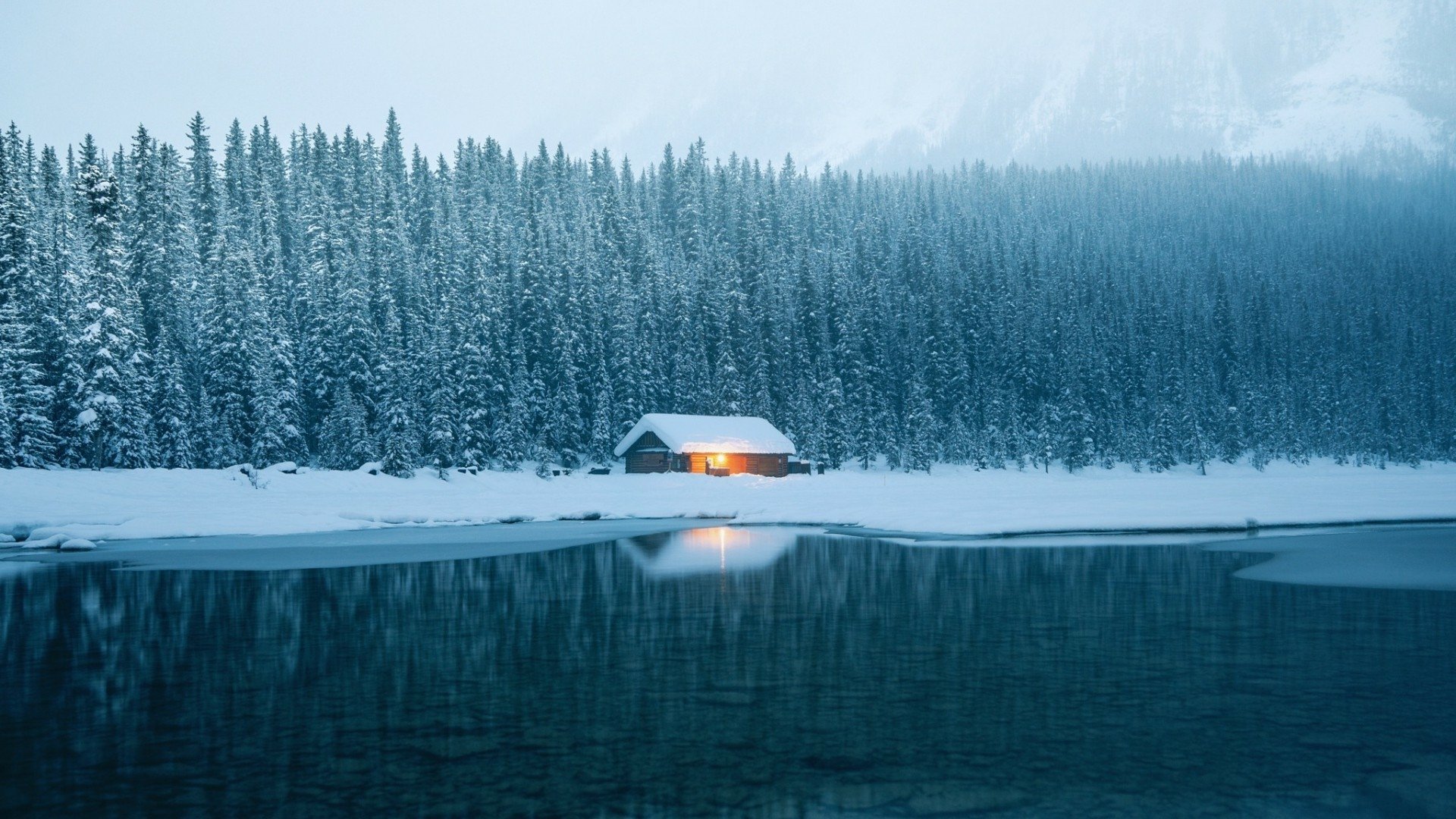 A serene winter scene featuring a snow-covered cabin by a calm lake, surrounded by tall evergreen trees, captured in HD for a desktop wallpaper background.