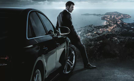 HD PC desktop wallpaper featuring a scene from the movie The Transporter Refueled, showing a man leaning against a black car overlooking a coastal city at dusk.