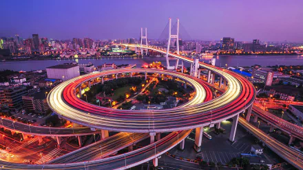 Time-lapse view of Nanpu Bridge’s circular highway ramp in Shanghai, China, showcasing vibrant cityscape and illuminated roads at twilight.
