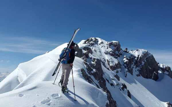 Mountaineer with skis ascending a snowy Alpine ridge beneath a clear winter sky — dramatic Alps mountain scene and mountaineering action. 4K Ultra HD PC desktop wallpaper.