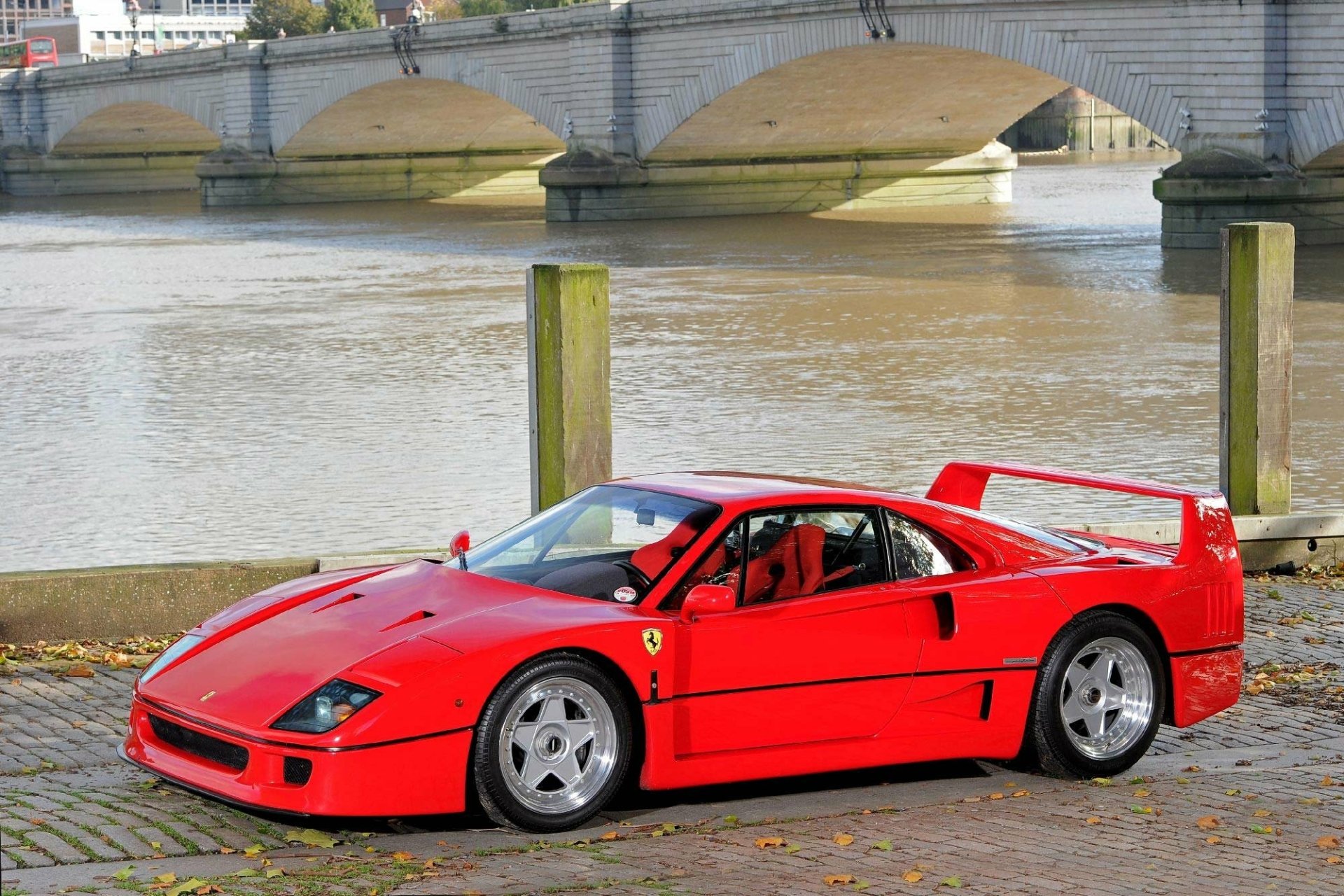 HD desktop wallpaper featuring a vibrant red Ferrari F40 parked by a riverside with a stone bridge in the background.
