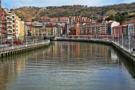 4K Ultra HD view of a man-made waterfront in Bilbao, Spain, showcasing urban buildings and cityscape along a calm river with hills in the background.