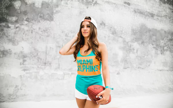 Brunette model wearing Miami Dolphins hat and tank top, holding a football against a textured concrete wall — 5K Ultra HD PC desktop wallpaper/background.