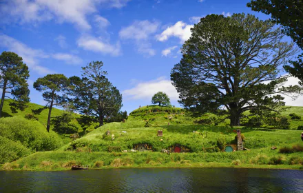 4K Ultra HD image of Hobbiton’s iconic man-made green hill homes nestled by a tranquil lake under a bright blue sky, surrounded by lush trees and greenery.