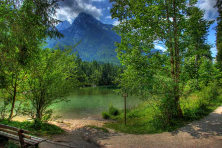 A serene landscape featuring a tranquil pond surrounded by lush green trees, a wooden bench, and towering mountains in the background, capturing the beauty of spring in Germany.