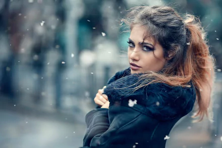 HD desktop wallpaper featuring a brunette woman with brown eyes, standing outdoors in a blurry background. The woman has a confident gaze and is wearing a dark coat, with light snow falling around her.
