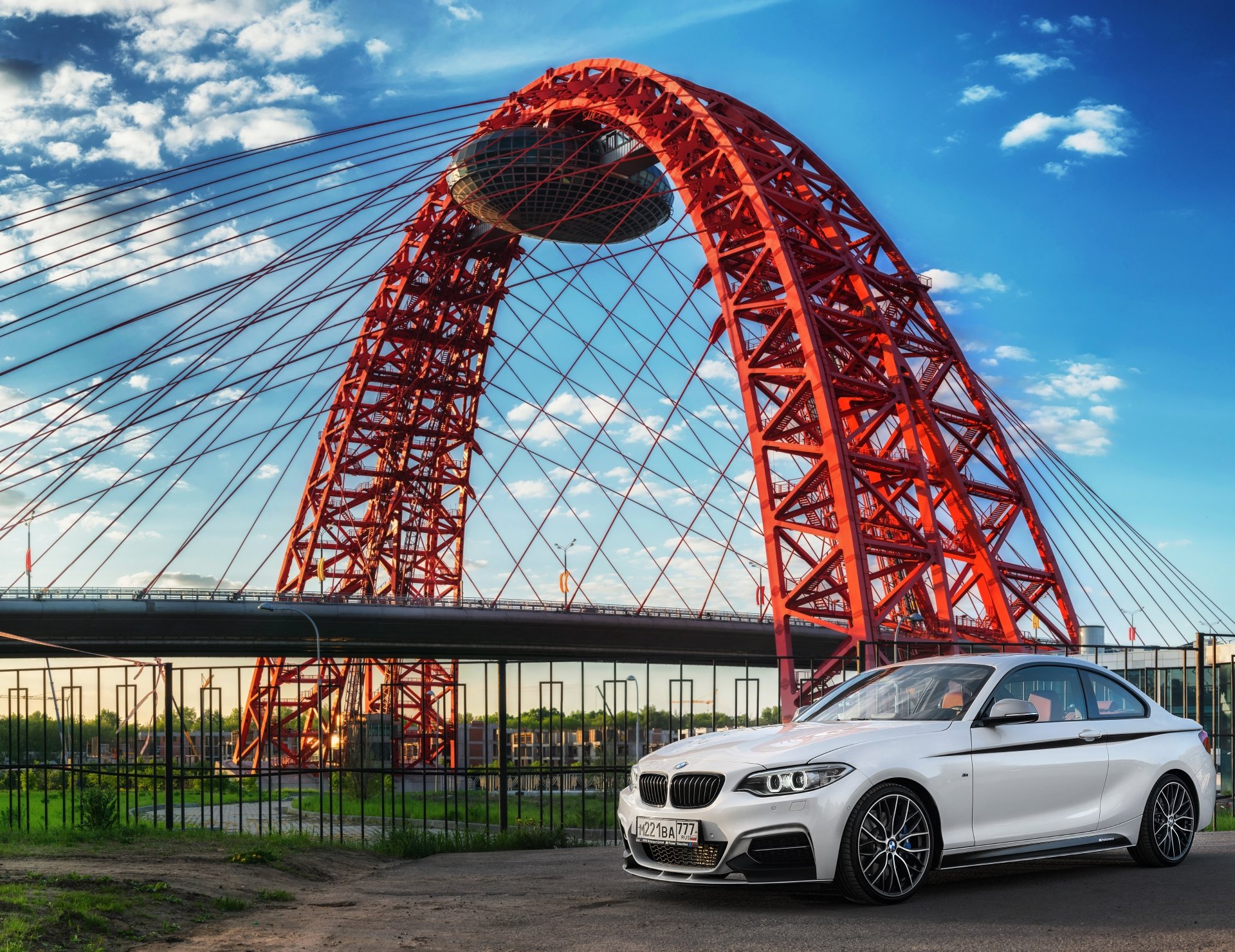 White BMW sedan parked by a riverside fence beneath a dramatic red arched bridge and blue sky — 5K Ultra HD PC desktop wallpaper and background.