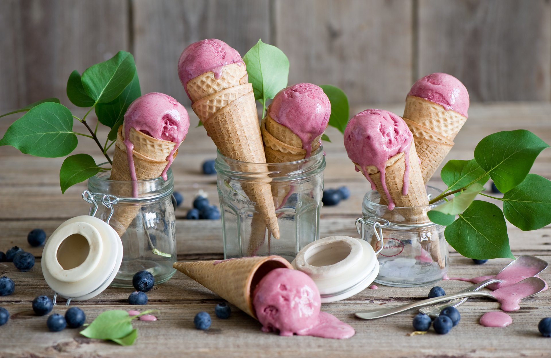 A vibrant still life featuring ice cream cones with blueberry-flavored ice cream, surrounded by fresh blueberries and green leaves, set against a rustic wooden background.