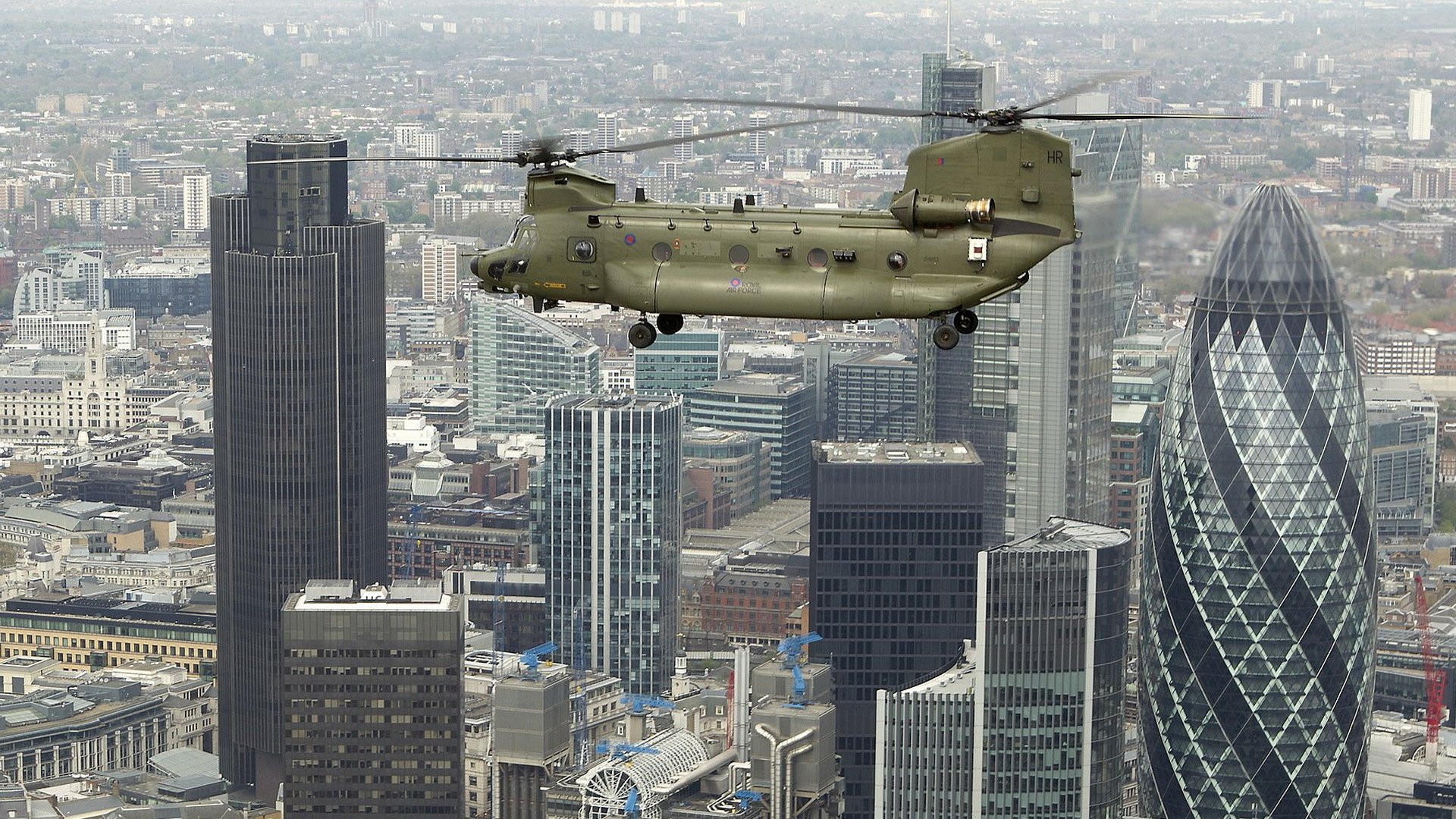 A military Boeing CH-47 Chinook transport helicopter flying over a city skyline, captured in high detail for an HD desktop wallpaper background.