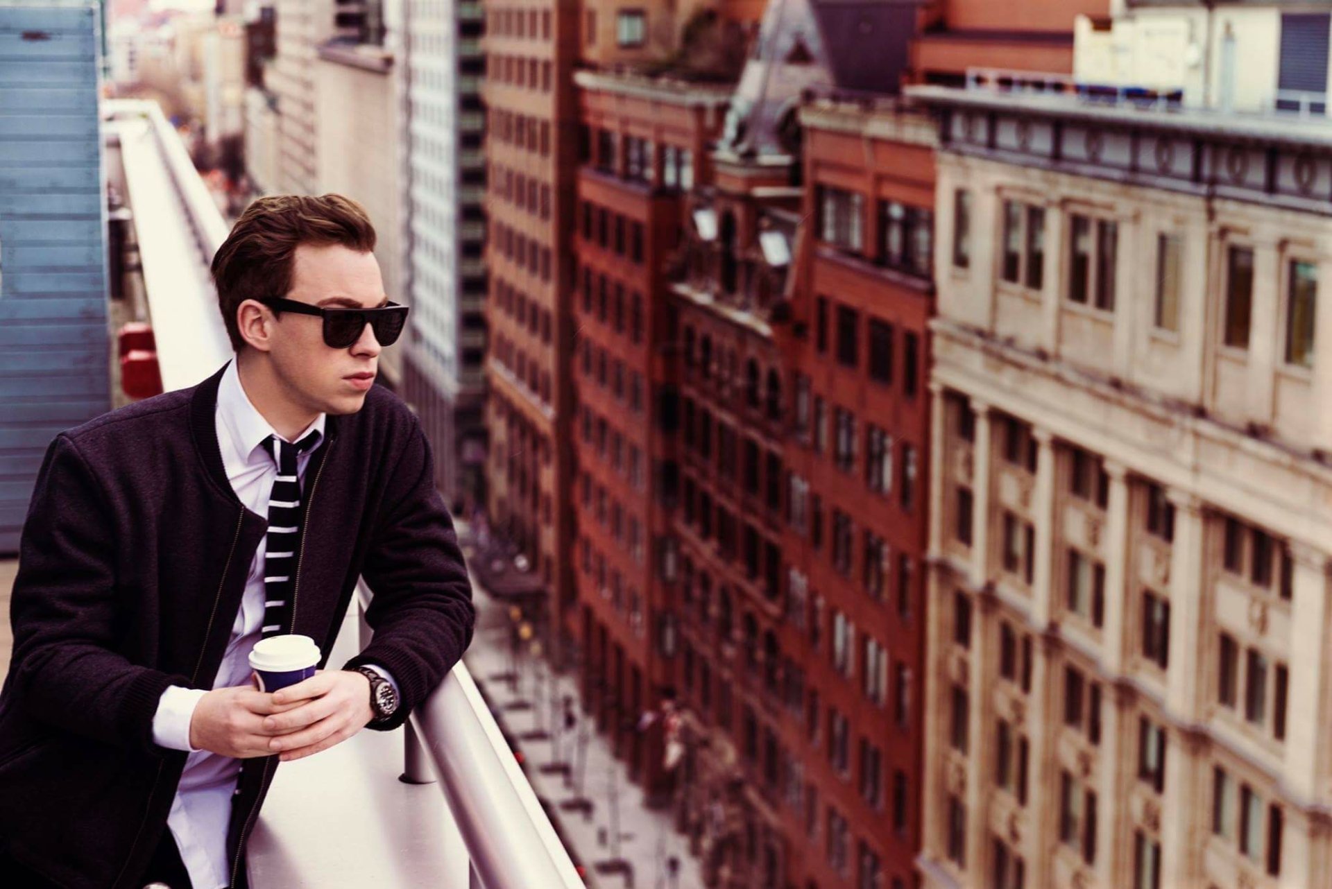 Young man with sunglasses holding coffee on a Montreal balcony overlooking the cityscape, capturing an urban vibe with music and a Hardwell-inspired mood.