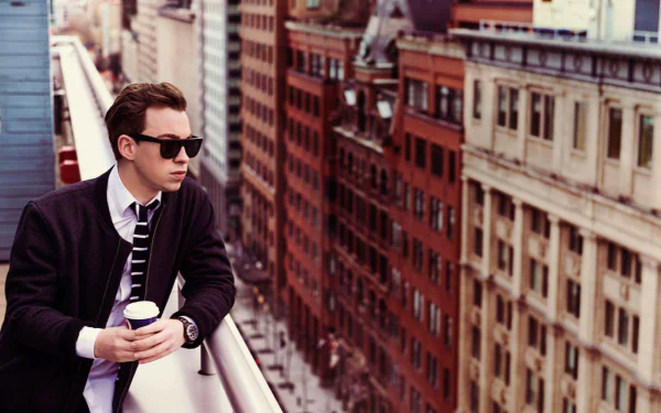 Young man with sunglasses holding coffee on a Montreal balcony overlooking the cityscape, capturing an urban vibe with music and a Hardwell-inspired mood.