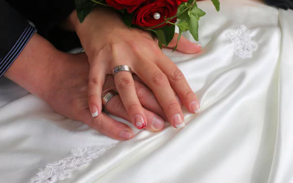 Close-up 4K Ultra HD image of intertwined hands wearing wedding rings, resting on a white wedding dress with a red rose bouquet, capturing love and wedding photography.