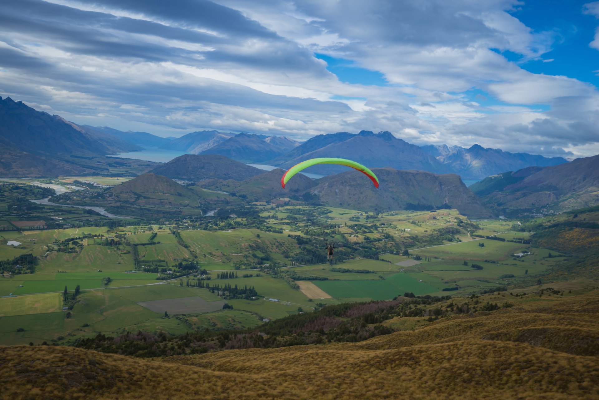 Paraglider soaring over a green patchwork valley and rugged mountains near Queenstown, New Zealand — 5K Ultra HD PC desktop landscape wallpaper and background.