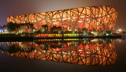 A stunning HD wallpaper featuring the iconic Bird's Nest stadium in Beijing, illuminated at night with vibrant orange lights, reflecting beautifully in the water below.