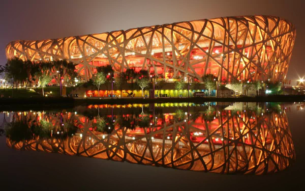 A stunning HD wallpaper featuring the iconic Bird's Nest stadium in Beijing, illuminated at night with vibrant orange lights, reflecting beautifully in the water below.