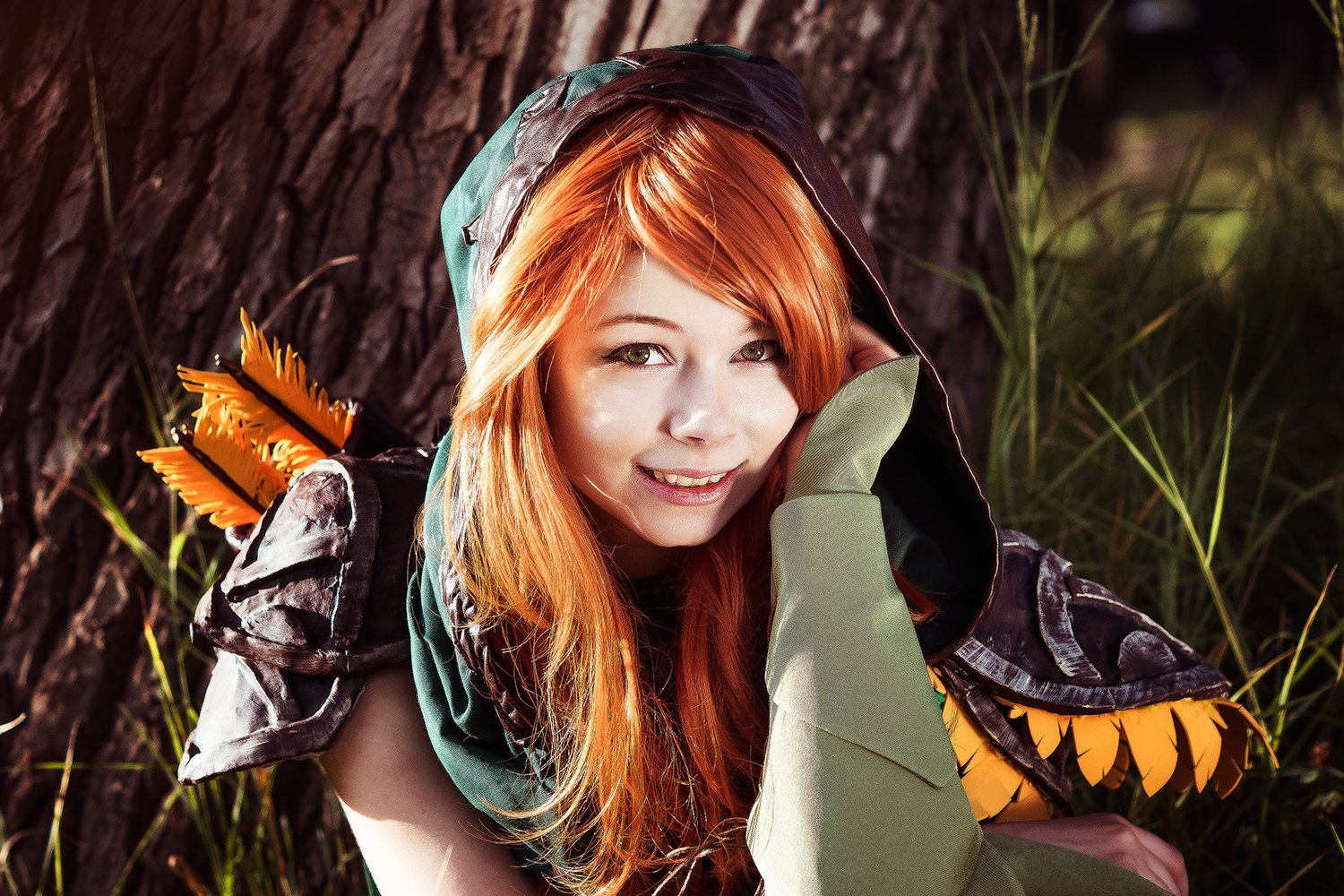 A woman in a Windranger cosplay from DotA 2 sits against a tree, smiling with her orange hair and vibrant green attire, creating a dynamic and engaging desktop wallpaper.