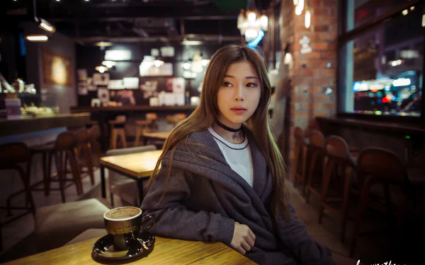 A brunette Asian woman with brown eyes sits thoughtfully at a pub table at night, with a cup of coffee in front of her, captured in 4K Ultra HD desktop wallpaper quality.
