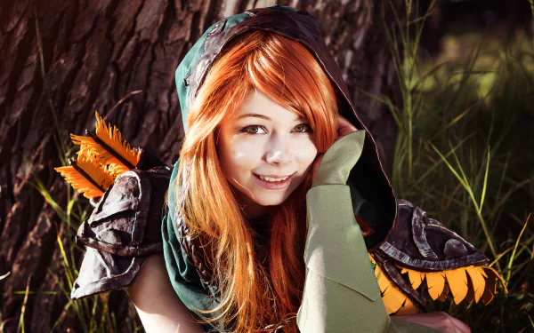 A woman in a Windranger cosplay from DotA 2 sits against a tree, smiling with her orange hair and vibrant green attire, creating a dynamic and engaging desktop wallpaper.