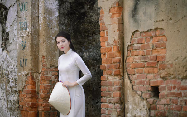 A Vietnamese model in a traditional white Ao Dai and Asian conical hat stands against a weathered brick wall, captured in stunning 4K Ultra HD detail.