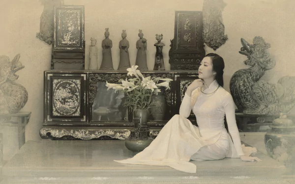 Sepia 5K Ultra HD desktop wallpaper of a Vietnamese Asian woman model in a white Ao Dai seated beside lilies before ornate antique cabinets and sculptures.