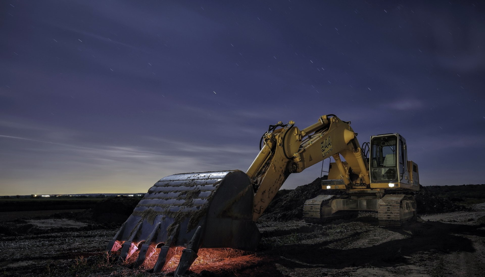 HD PC desktop wallpaper of a yellow excavator at night, its bucket casting a red glow on dark soil beneath a star-filled sky