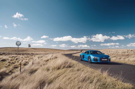A striking Audi R8 V10 sports car drives along a scenic country road, surrounded by open fields and a clear blue sky, showcasing its sleek design in this 4K Ultra HD wallpaper.