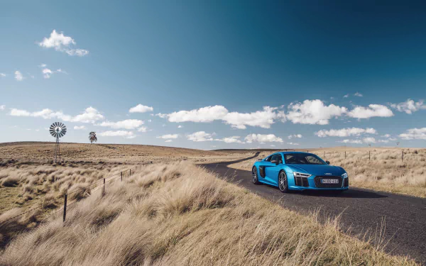 A striking Audi R8 V10 sports car drives along a scenic country road, surrounded by open fields and a clear blue sky, showcasing its sleek design in this 4K Ultra HD wallpaper.