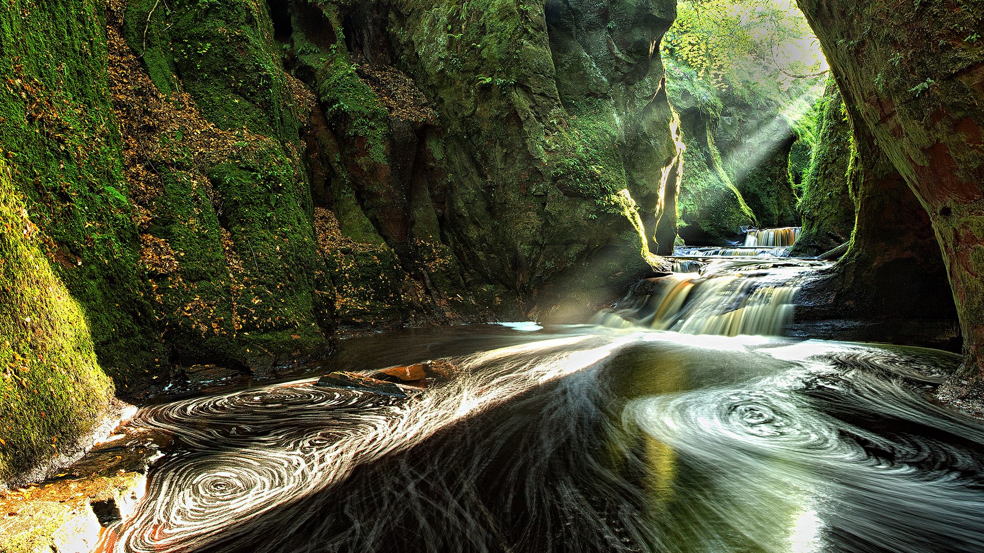 An HD desktop wallpaper showing a stunning canyon in Scotland with a cascading waterfall and sunbeams illuminating the lush green nature within a cave-like setting.