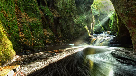 An HD desktop wallpaper showing a stunning canyon in Scotland with a cascading waterfall and sunbeams illuminating the lush green nature within a cave-like setting.