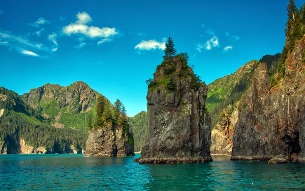 HD desktop wallpaper showcasing a serene Alaska fjord with towering cliffs and rocky formations rising from calm waters under a bright blue sky.