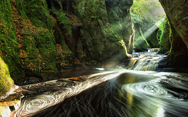An HD desktop wallpaper showing a stunning canyon in Scotland with a cascading waterfall and sunbeams illuminating the lush green nature within a cave-like setting.
