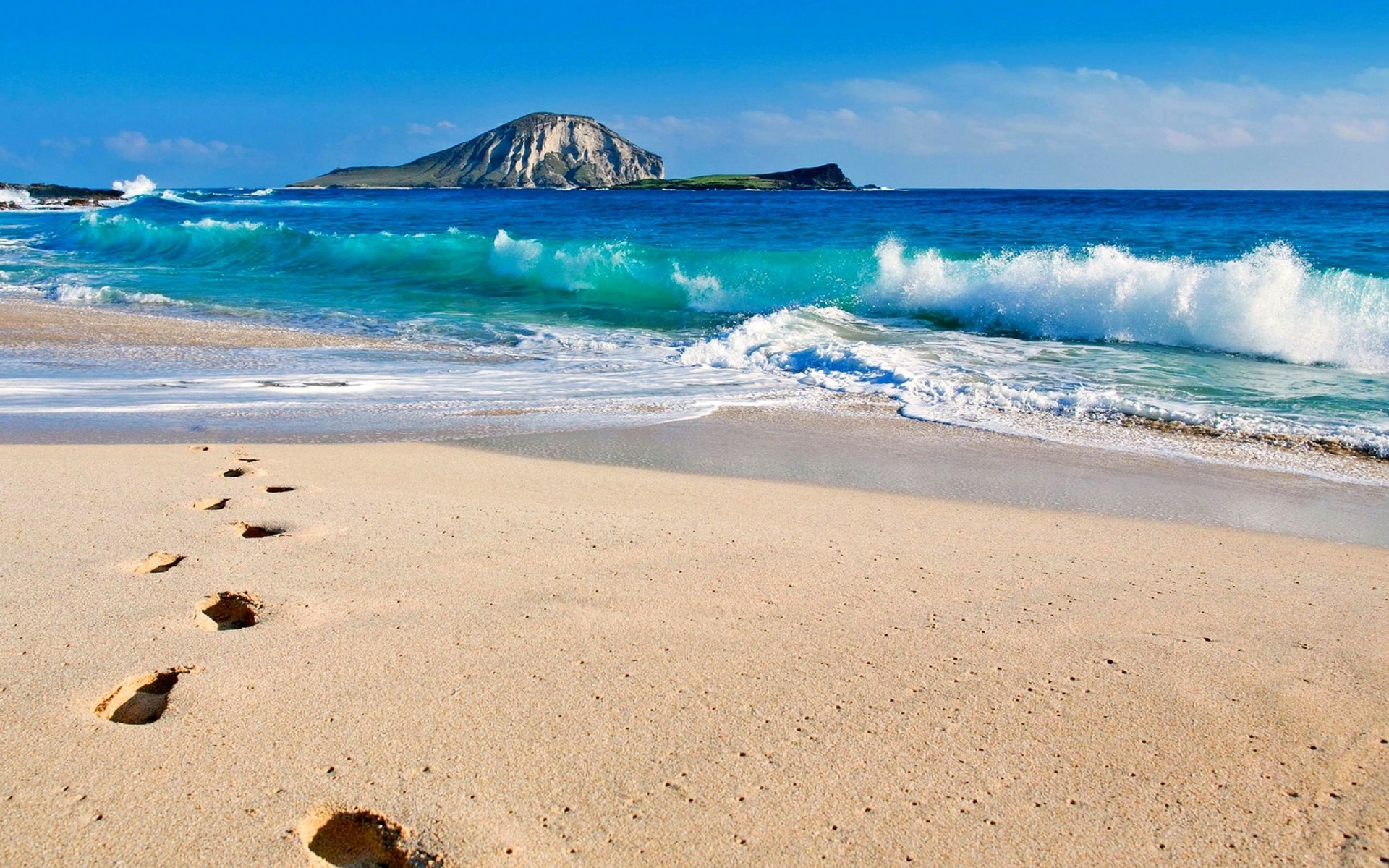 Footprints trail across a sandy beach with gentle waves rolling in from the ocean toward a distant island under a clear blue horizon in this HD desktop wallpaper.