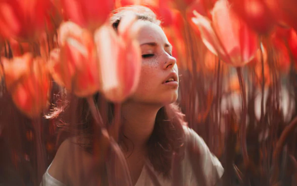 A brunette woman with freckles among red flowers, eyes closed, immersed in a serene mood. HD PC desktop wallpaper and background.