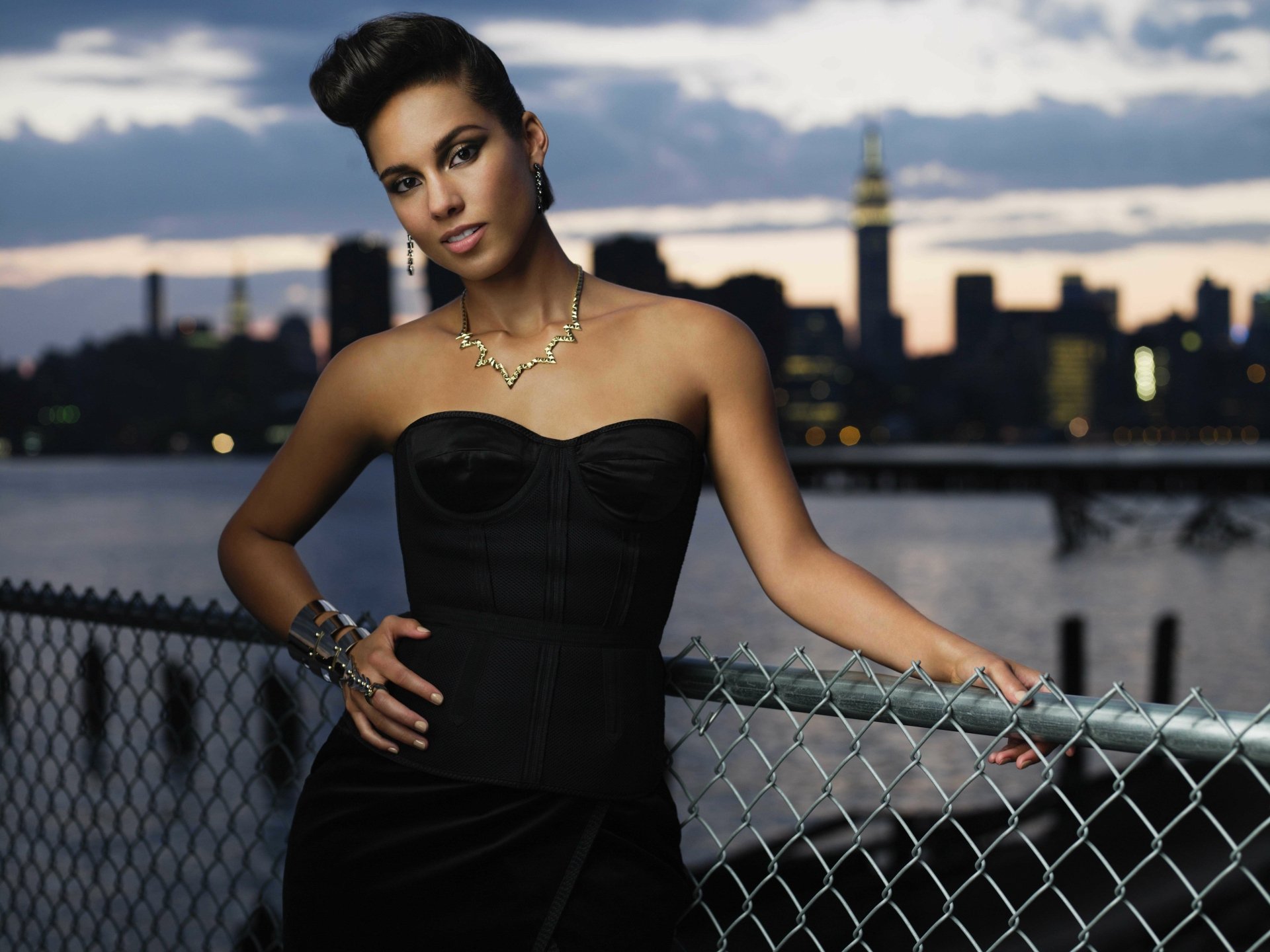Brunette woman with brown eyes wearing a necklace and earrings, posed by an urban riverside skyline at dusk — 5K Ultra HD desktop wallpaper with a musical performer vibe.