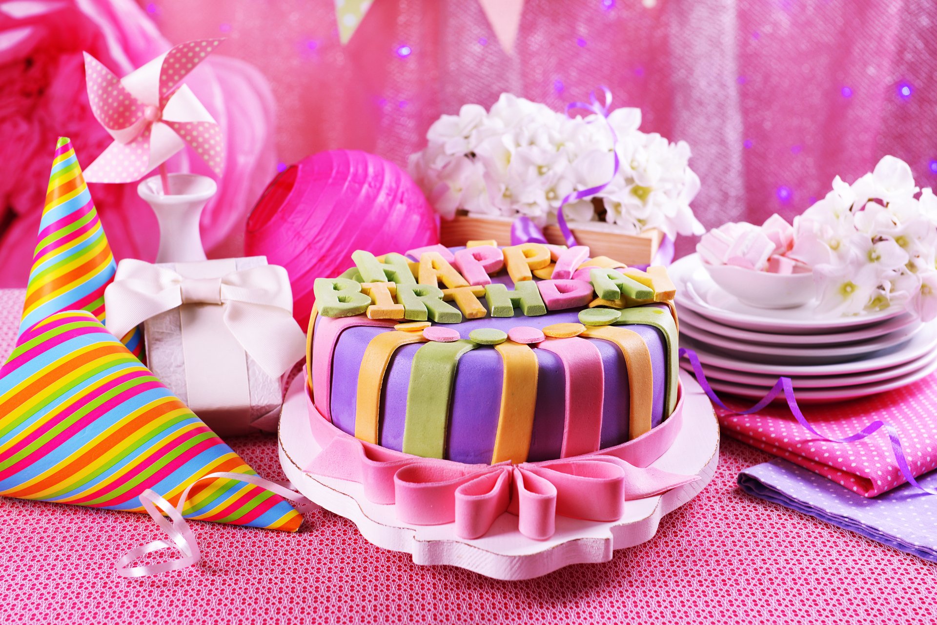 A vibrant birthday cake with colorful icing sits among pink decorations, gifts, and flowers, all set against a festive backdrop, creating a joyful holiday atmosphere.