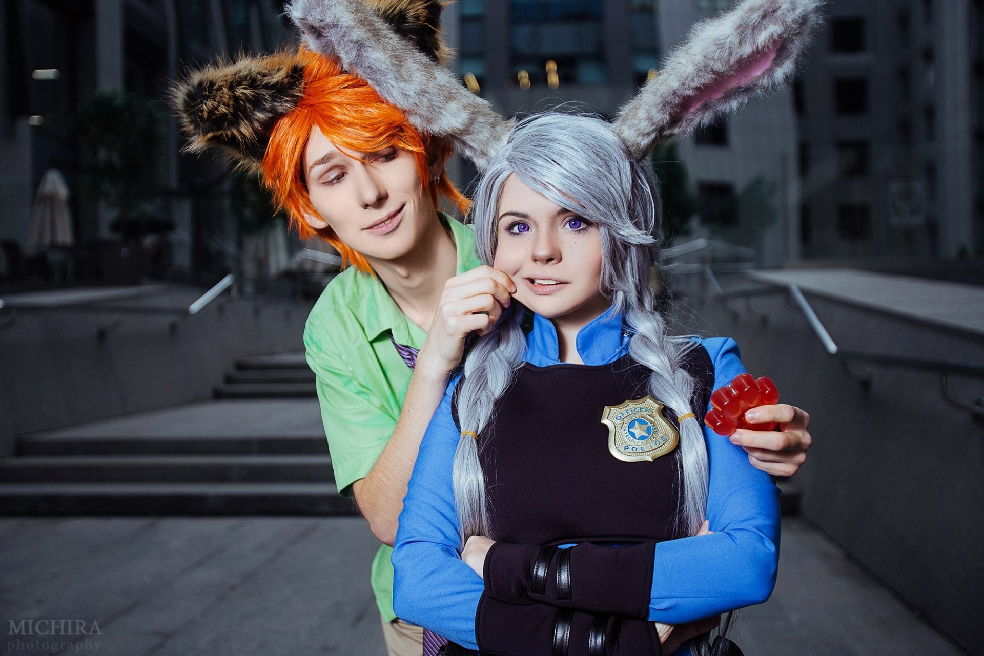 HD PC desktop wallpaper: two women cosplaying Judy Hopps and Nick Wilde from Zootopia — Judy in a blue uniform with bunny ears and badge, Nick in a green shirt with fox ears holding a red treat.